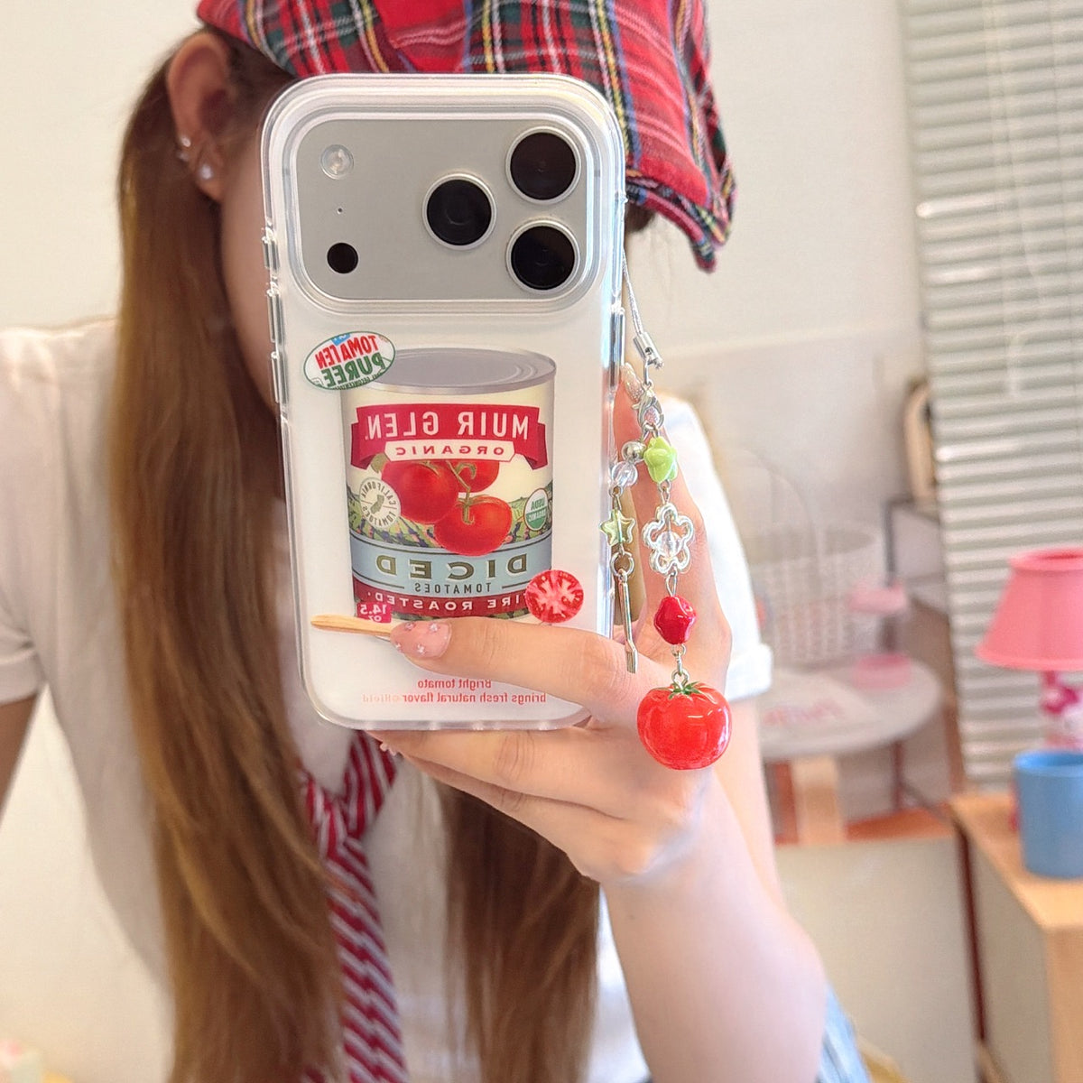 A girl taking a mirror selfie holding a retro tomato can iPhone case with a cute tomato charm lanyard.