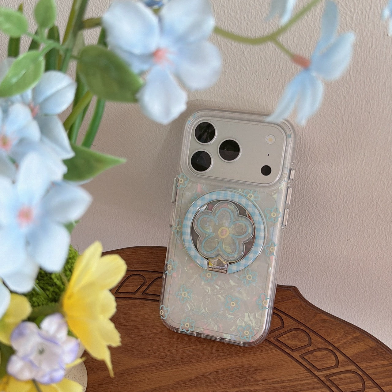 A clear blue flower pearl iPhone case standing on a wooden table behind beautiful blurred light-blue blossoms.