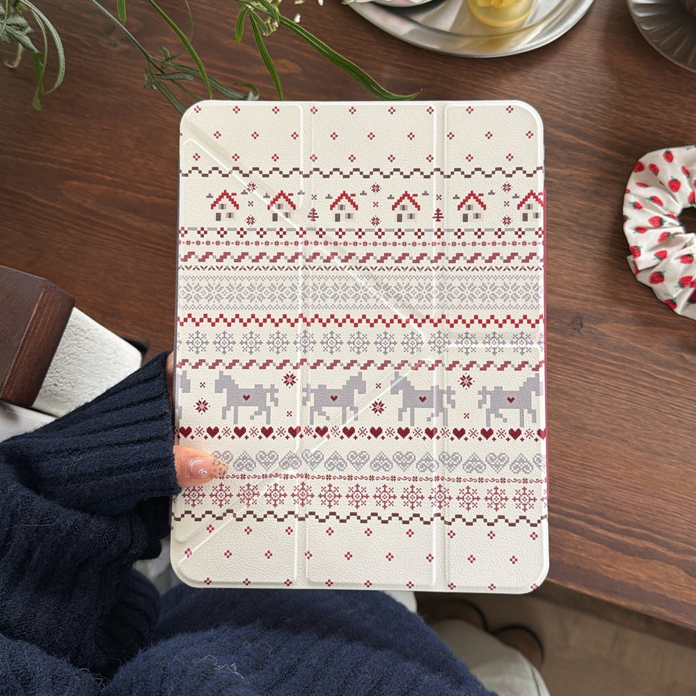 A hand holding a scandinavian pattern iPad smart cover showing pixel art houses and horses over a dark wooden desktop next to a strawberry scrunchie.