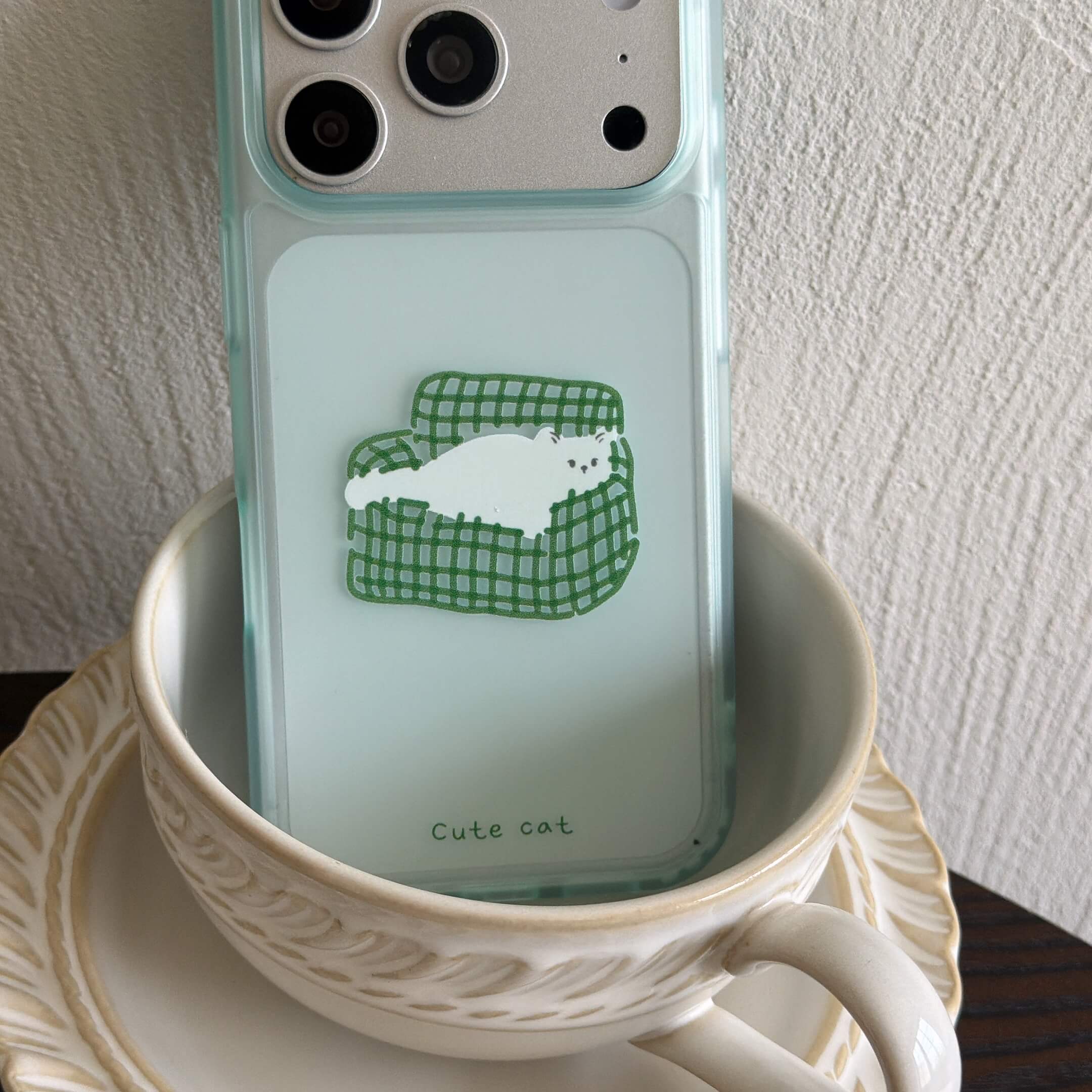 A shockproof green-rimmed cat graphic iPhone case standing inside an elegant white teacup.