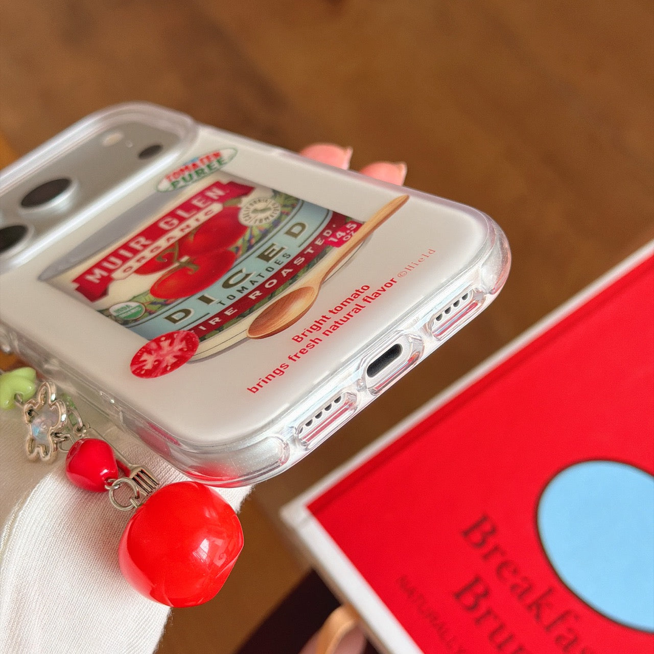 Close-up view of the shockproof TPU bumper edge, charging port, and dangling red tomato pendant of the case.
