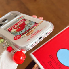 Close-up view of the shockproof TPU bumper edge, charging port, and dangling red tomato pendant of the case.