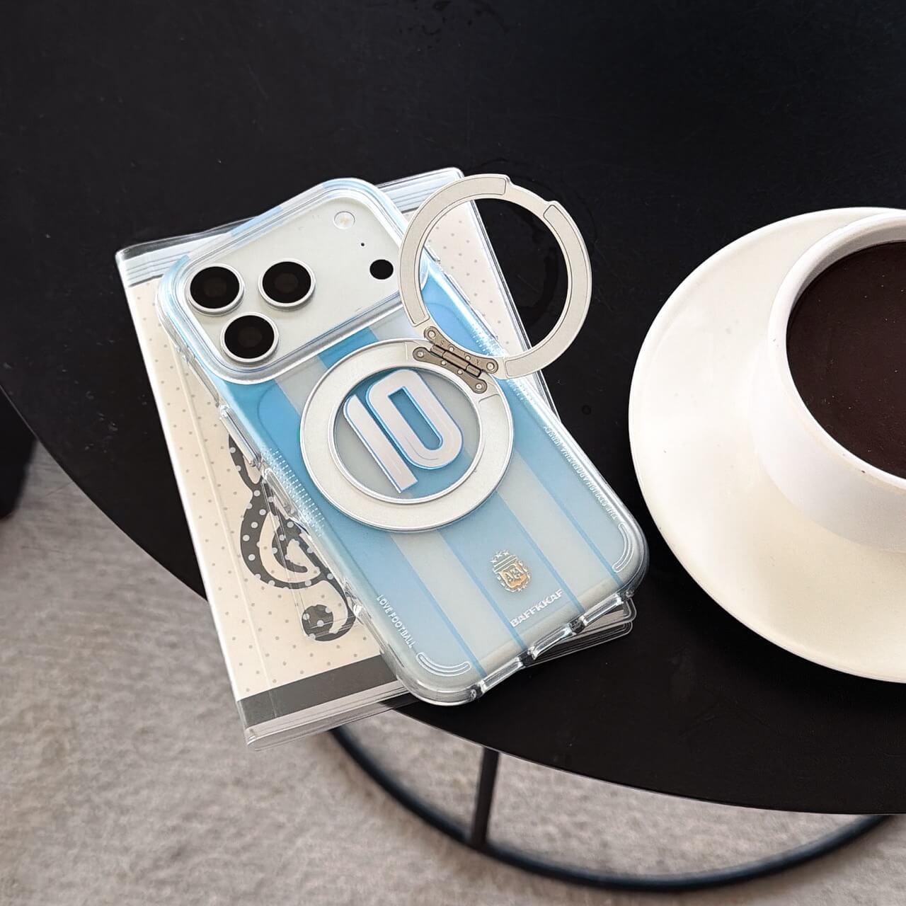 A soccer jersey iPhone case with its silver ring stand opened next to a white coffee cup.