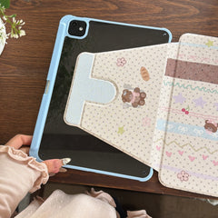 Close-up of a hand unfolding the smart magnetic cover of a pastel blue iPad case, revealing the cute teddy bear illustration on a polka-dot background over a rich wood grain surface.