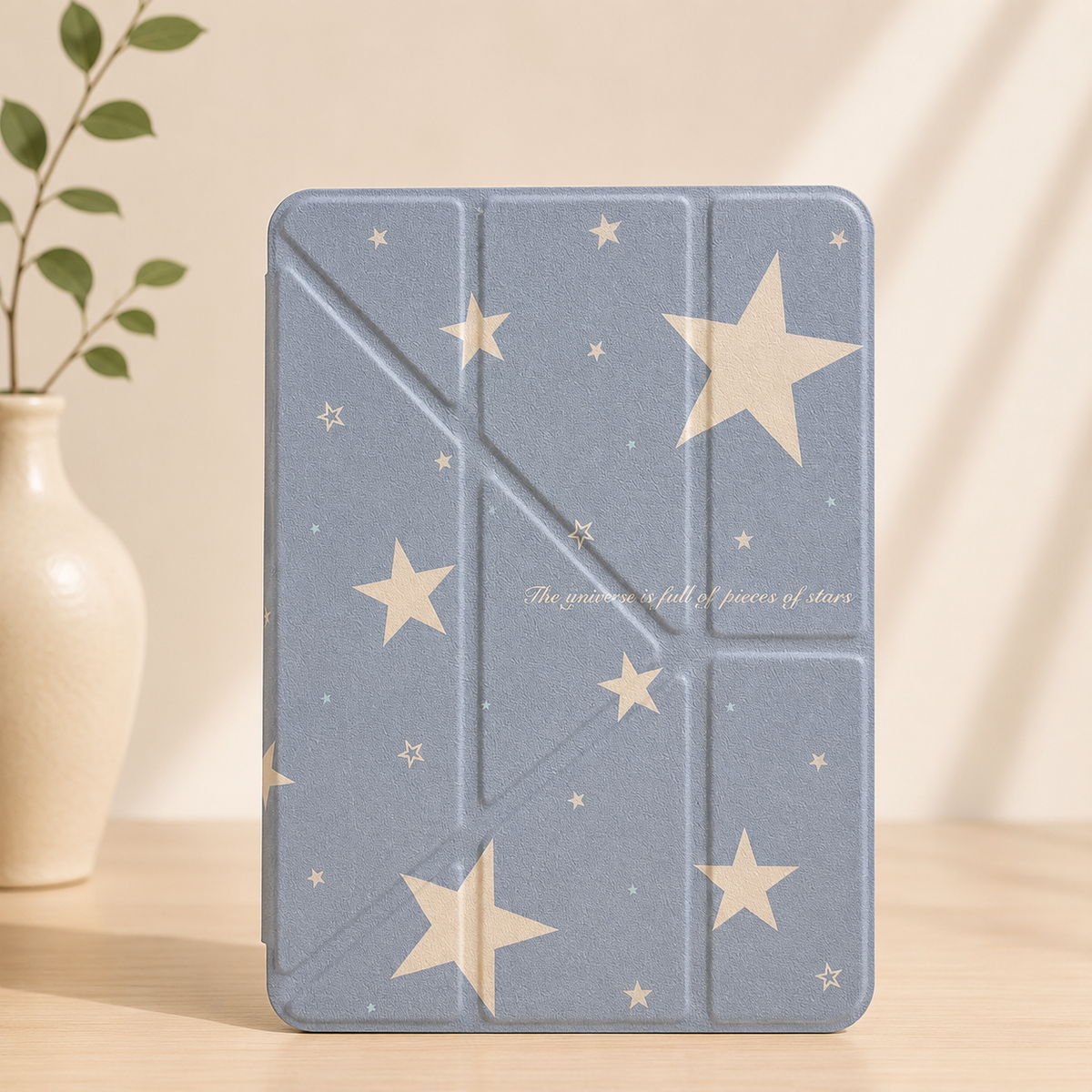 A pastel blue starry sky aesthetic origami iPad case standing vertically on a wooden table next to a white ceramic vase with green leaves.