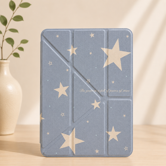 A pastel blue starry sky aesthetic origami iPad case standing vertically on a wooden table next to a white ceramic vase with green leaves.