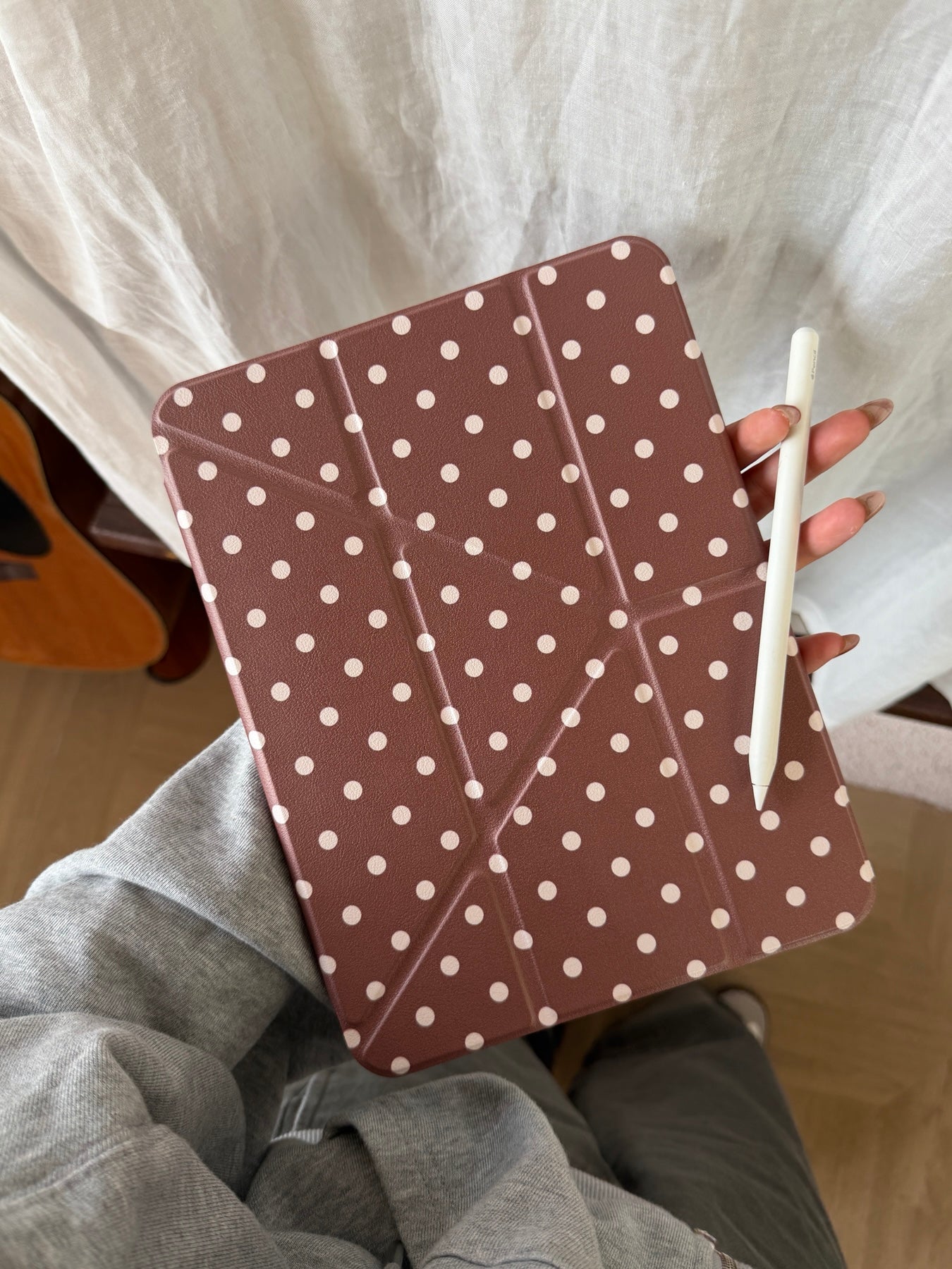 A hand holding a taupe brown polka dot ipad case with geometric folding lines next to a white stylus pen against a neutral fabric background.