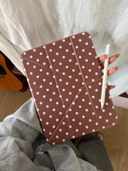A hand holding a taupe brown polka dot ipad case with geometric folding lines next to a white stylus pen against a neutral fabric background.