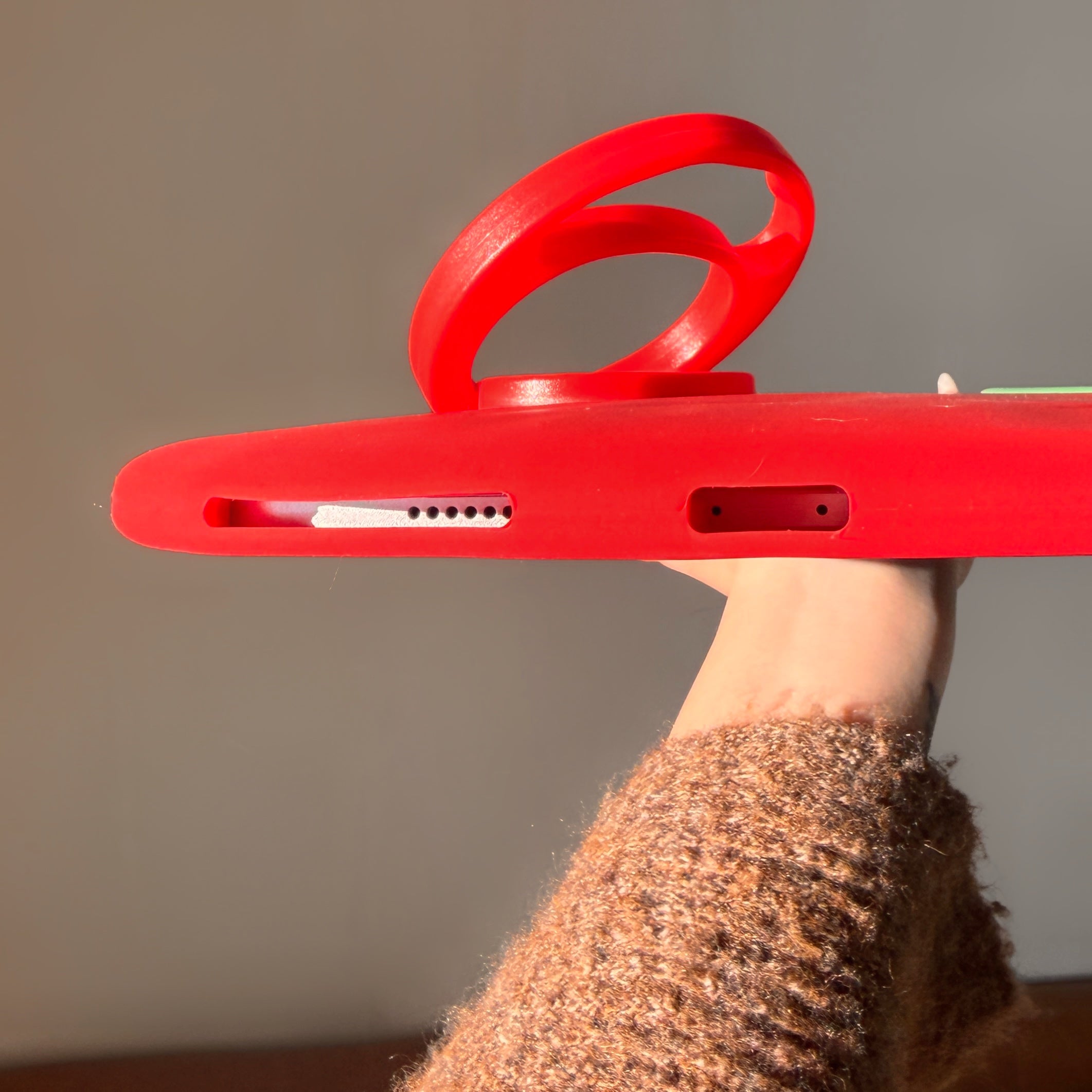 Close-up side view of a tomato tablet case with ring holder, highlighting the precise speaker holes and charging port cutout on the bottom silicone edge as a hand holds it up.