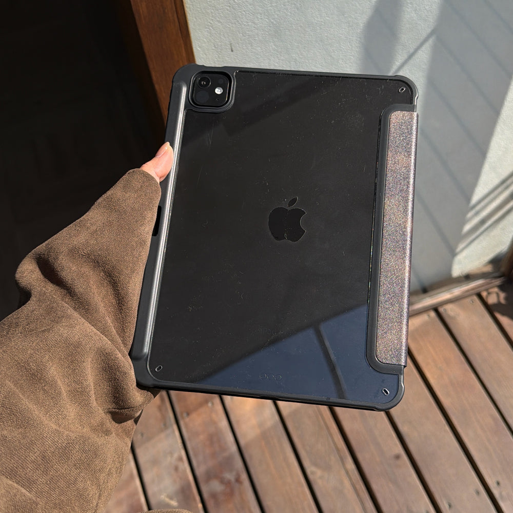 A hand holding the translucent black matte back shell of a tortoiseshell ipad case in the sunlight, showcasing the Apple logo and protective bumper edges over a wooden deck.