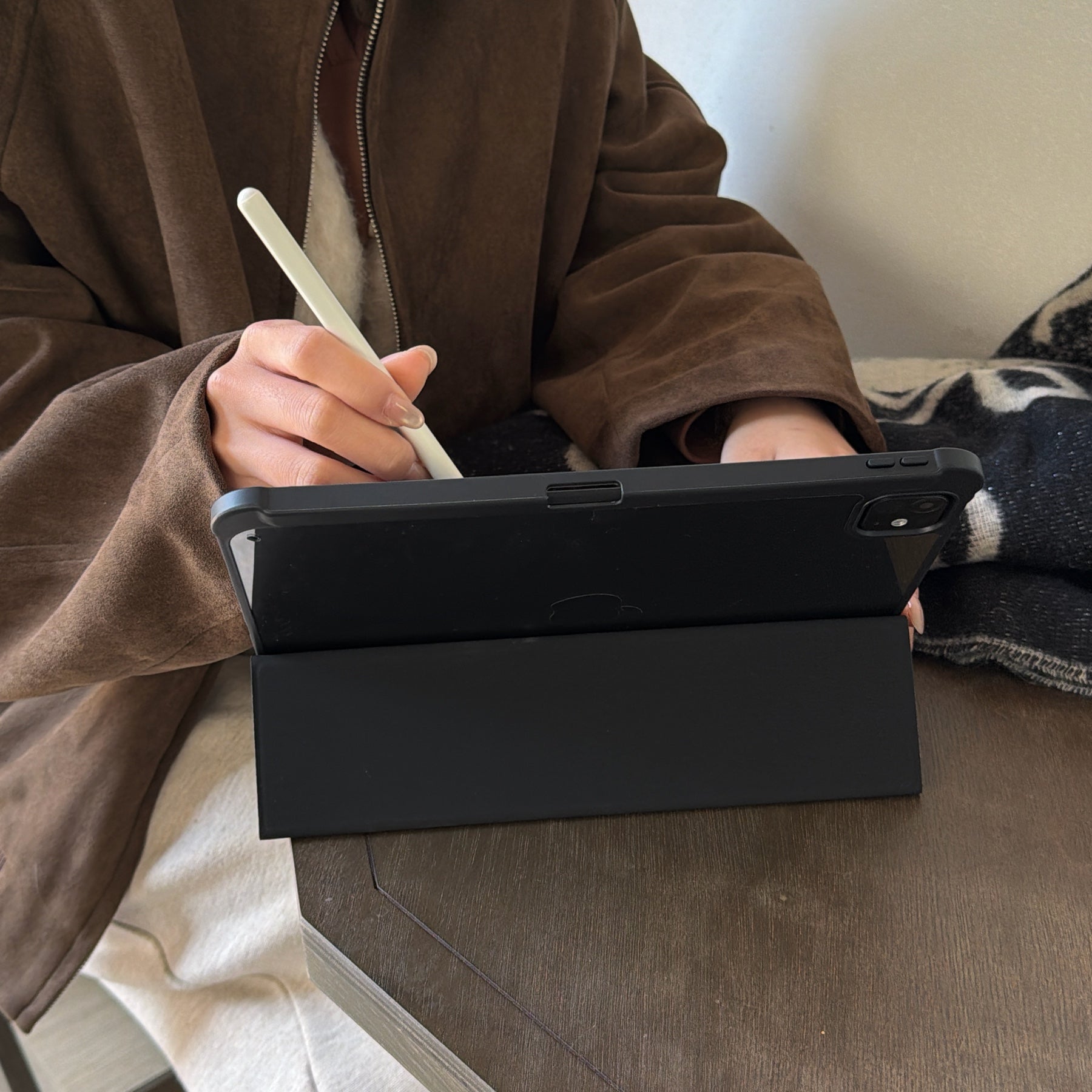 A user sketching with a white stylus on an iPad supported by a black tri-fold smart cover case stand, back angle view showing the sleek matte finish.