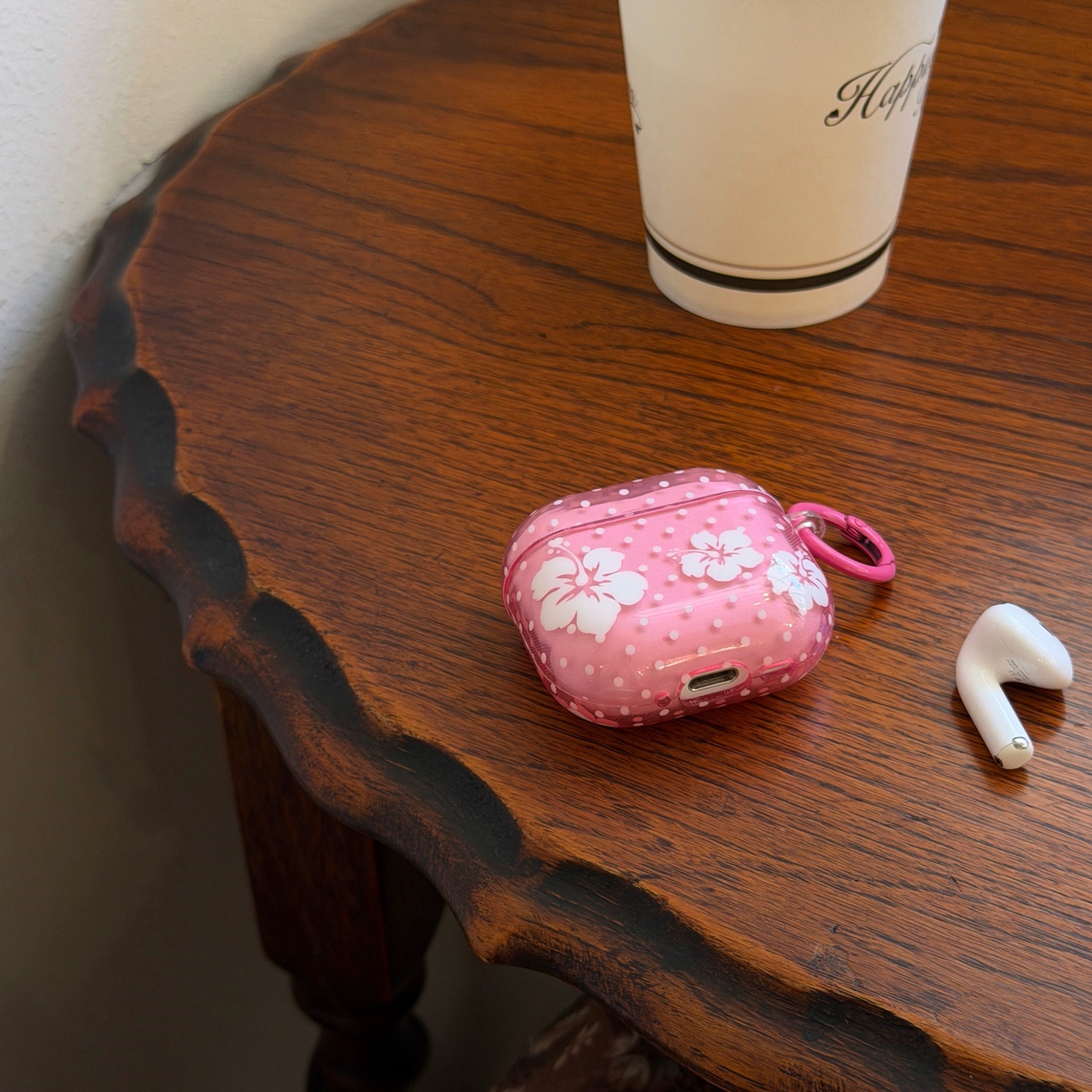 A tropical pink flower AirPods case and a single wireless earbud resting on the edge of a vintage wooden table.