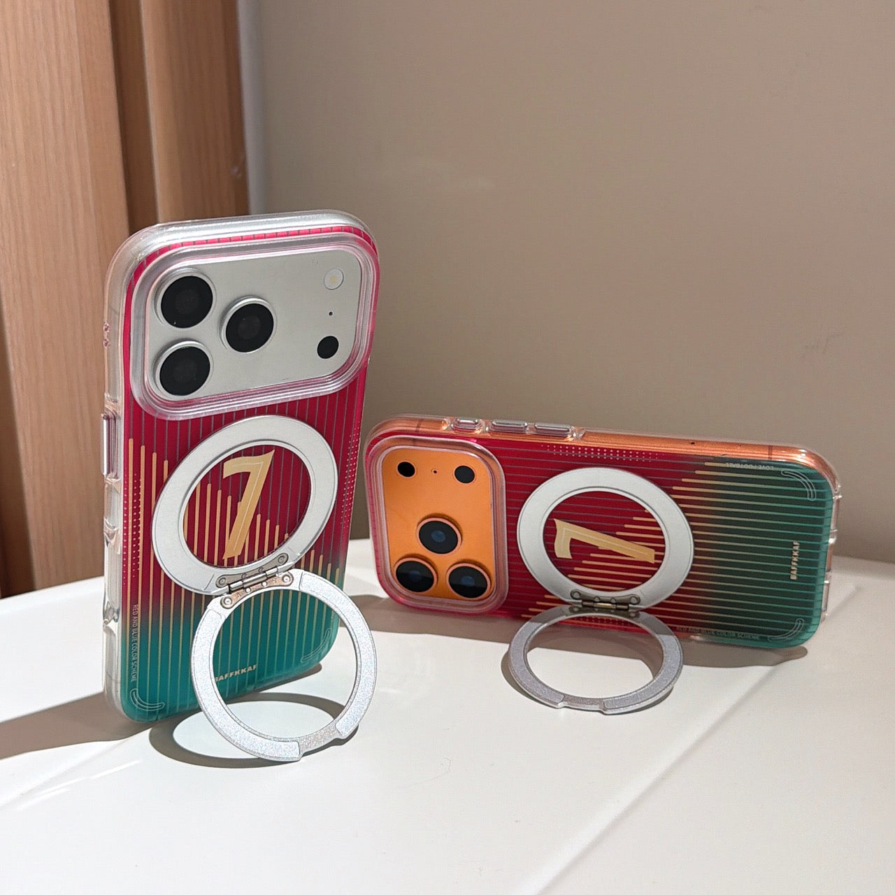 Two iphones, one white and one orange, standing on a white surface using the built-in silver magnetic ring kickstands of their matching red and teal gradient covers.