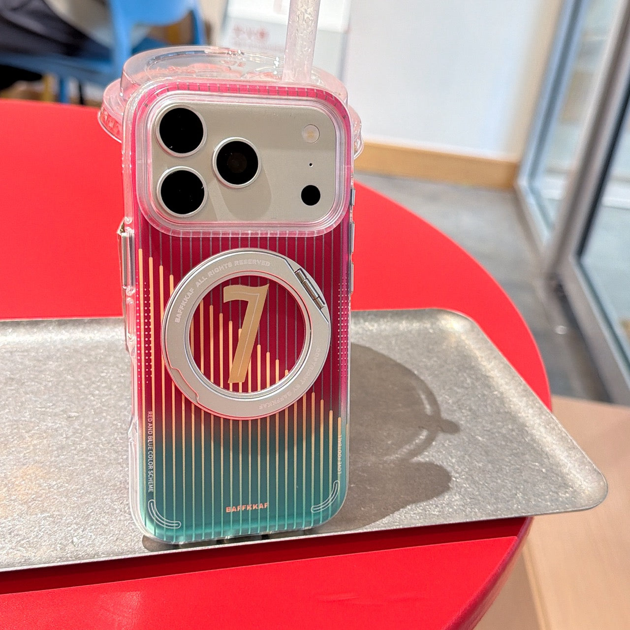 A transparent mobile phone cover with a sporty red and green gradient and number 7 print, standing upright against a plastic cup on a silver tray.