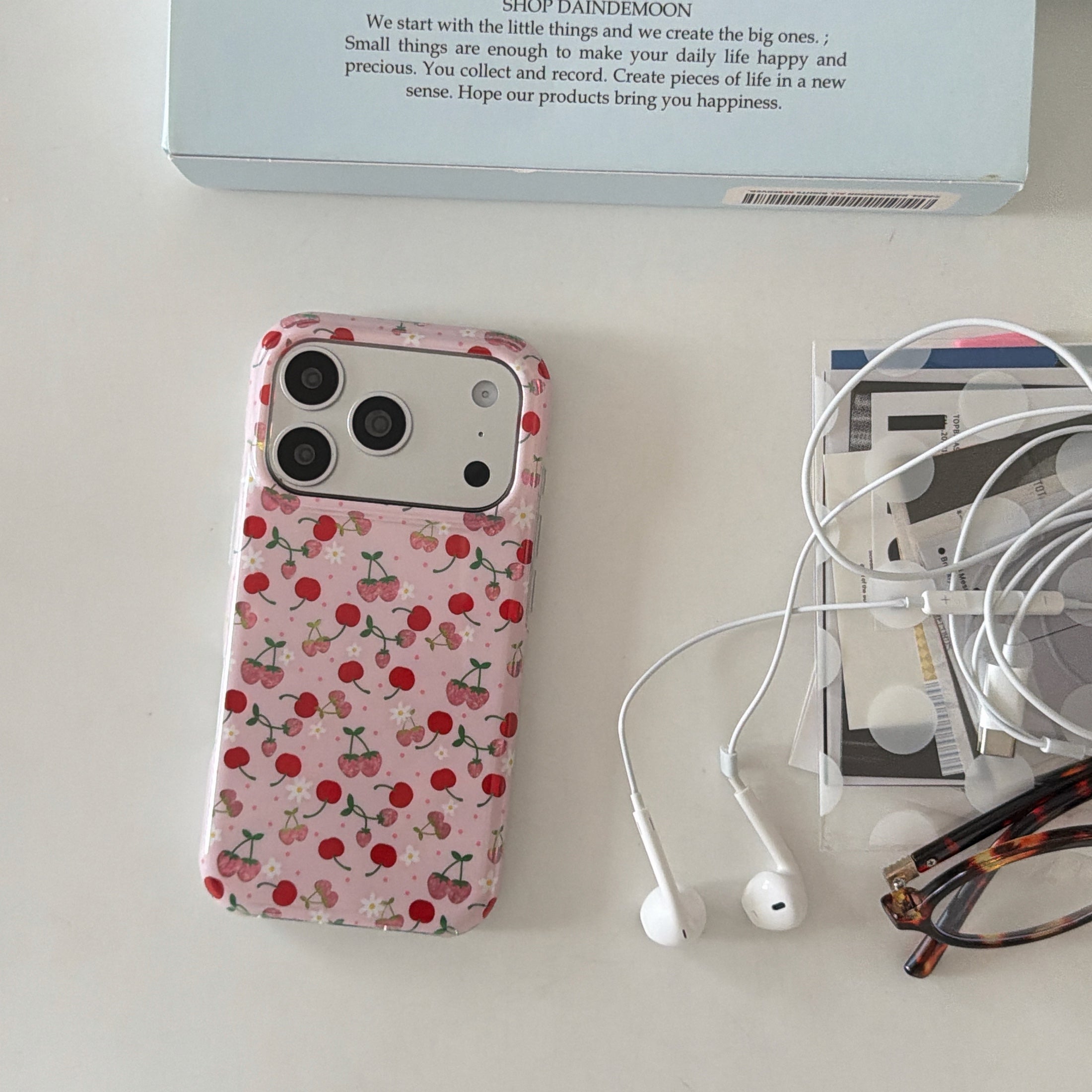A vintage cherry phone case on a desk next to white earphones, reading glasses, and a blue box.