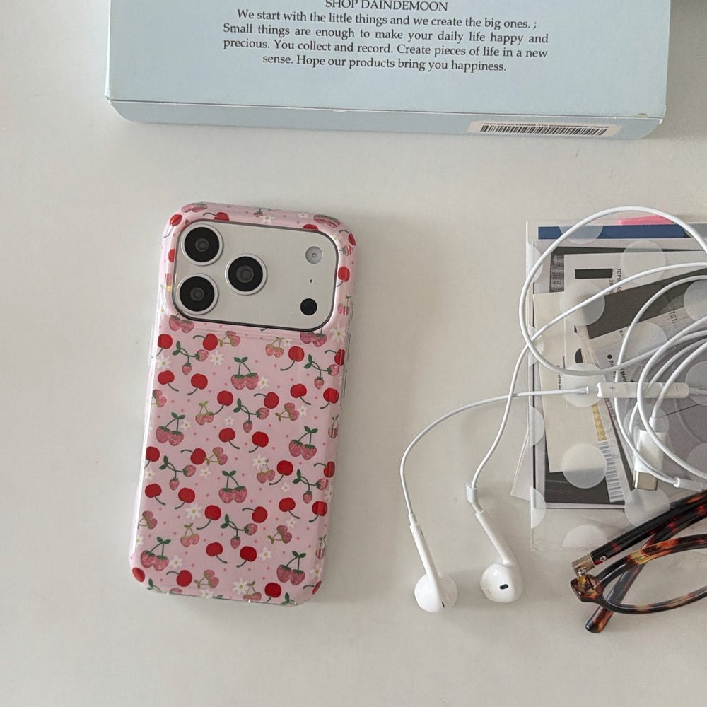 A vintage cherry phone case on a desk next to white earphones, reading glasses, and a blue box.