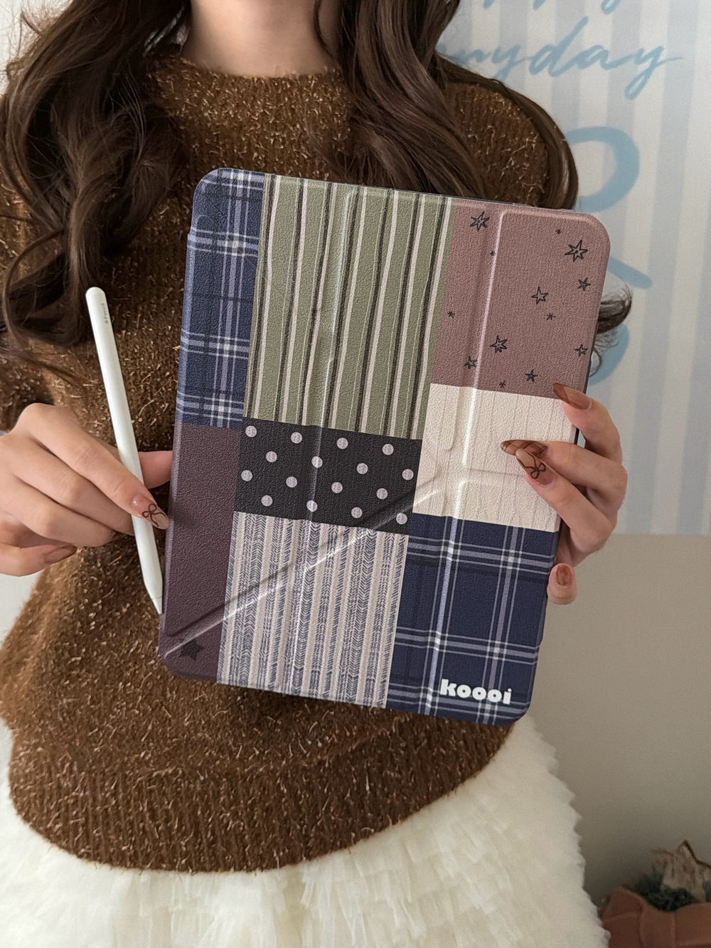 A model wearing a cozy brown sweater holding the vintage dark academia style patchwork iPad case vertically alongside a white Apple pencil.