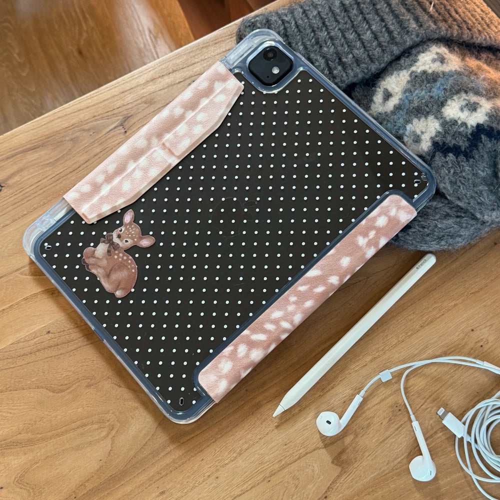 A flat lay showcasing the back of a black polka dot iPad protective shell featuring a vintage deer illustration, an Apple Pencil, and earphones on a wooden desktop.