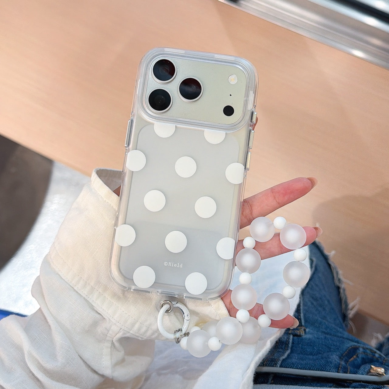 A hand holding a white polka dot clear iPhone case featuring a frosted white beaded wrist strap.