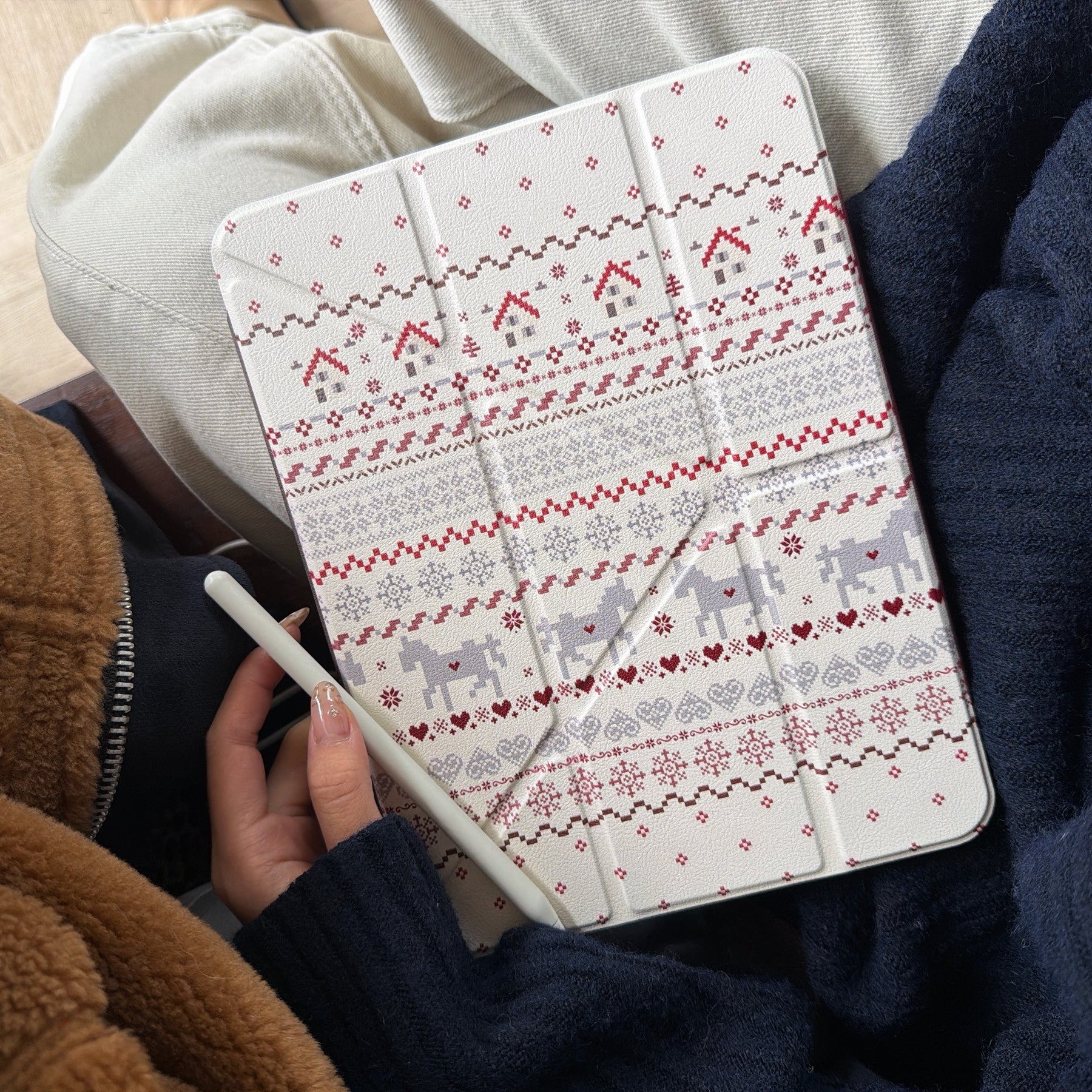 A close-up view of a person wearing a cozy winter sweater holding a Nordic-patterned winter aesthetic iPad cover while holding a white stylus pencil in their hand.