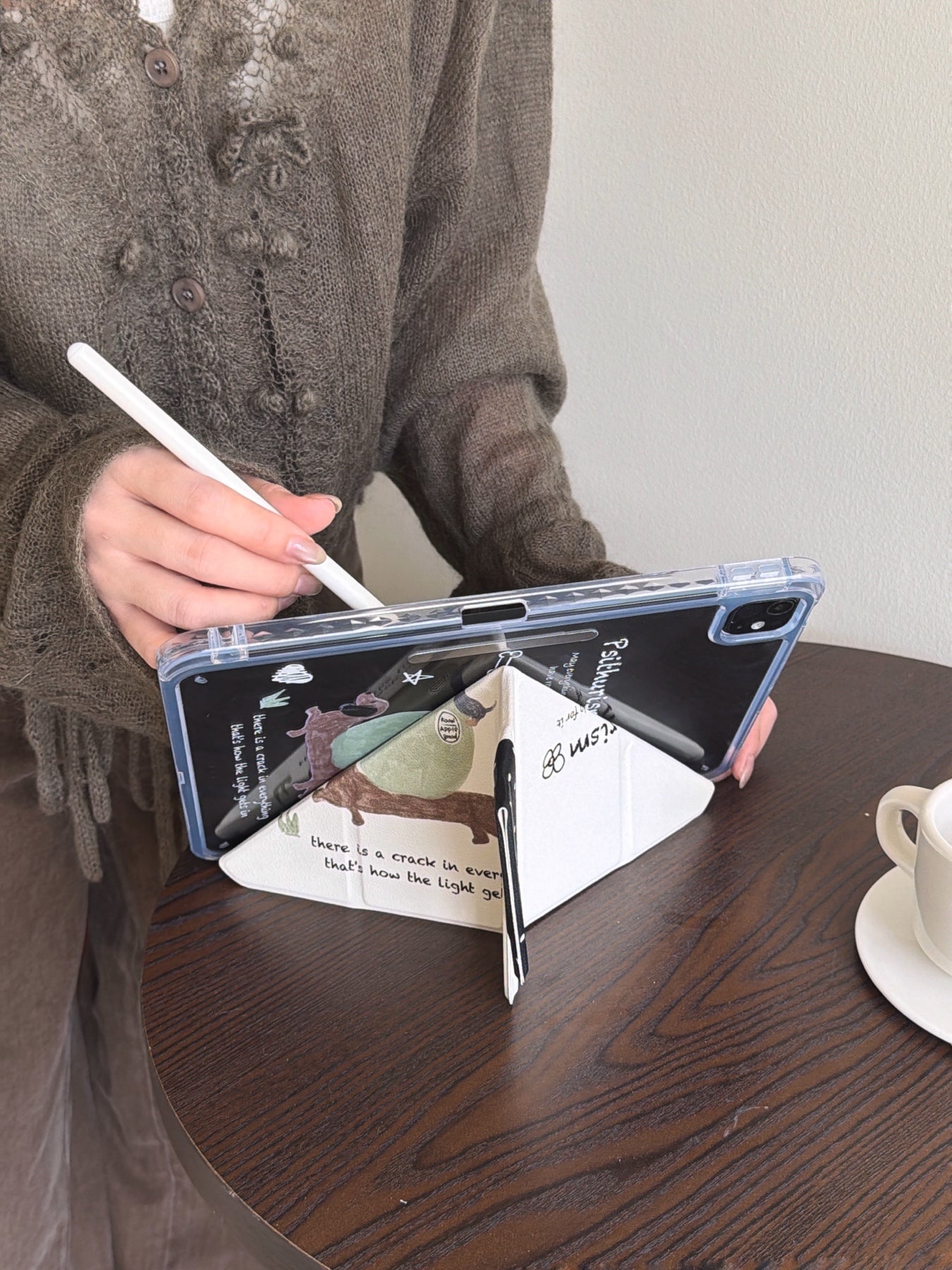 A close-up of a y fold trifold ipad case with pencil holder folded into a triangular stand on a dark wooden cafe table, with a hand holding a white stylus nearby.
