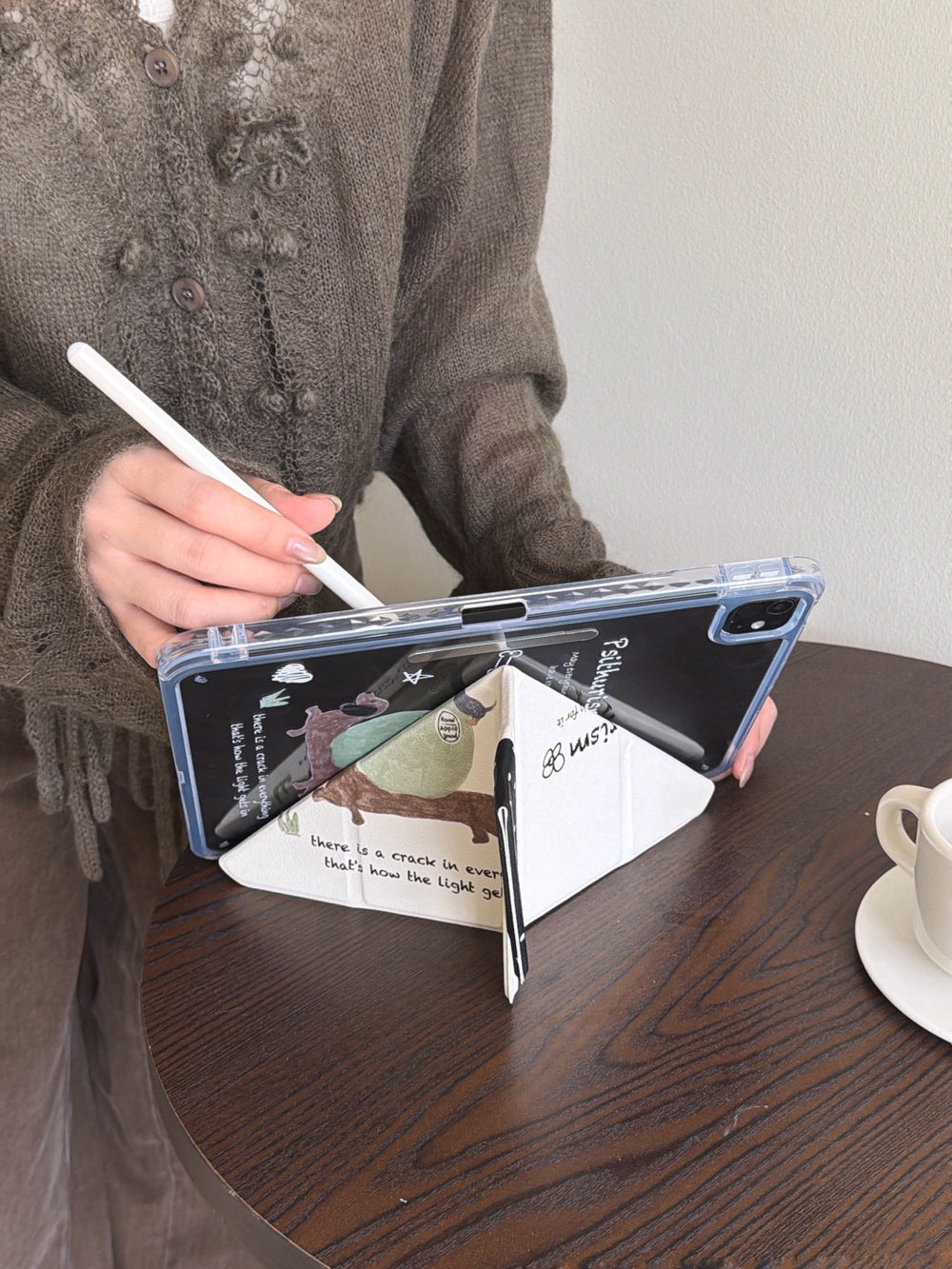 A close-up of a y fold trifold ipad case with pencil holder folded into a triangular stand on a dark wooden cafe table, with a hand holding a white stylus nearby.