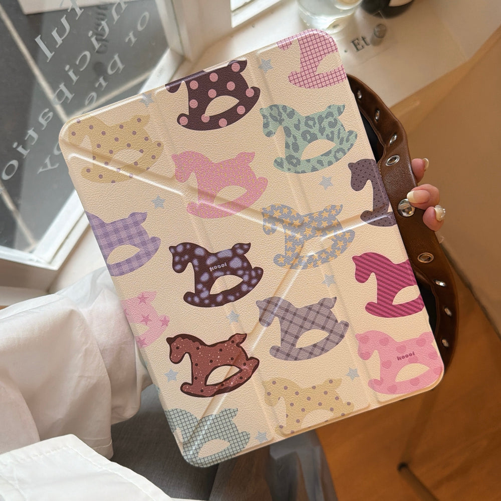 A hand holding a closed y2k aesthetic clear back ipad folio featuring a colorful pastel rocking horse design near a window.