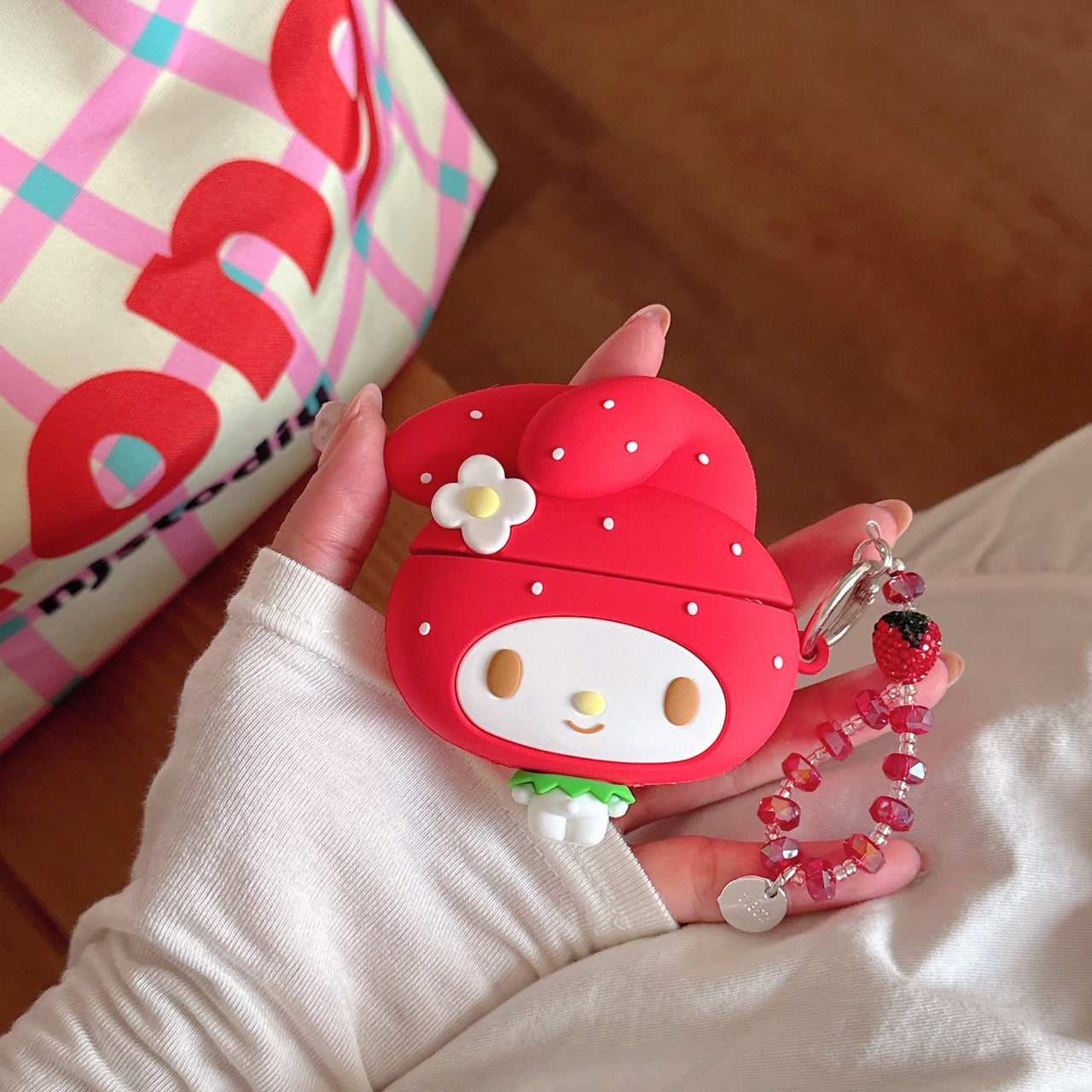 A hand holding a 3D cartoon strawberry style AirPods protective shell with a red crystal charm against a checkered bag.