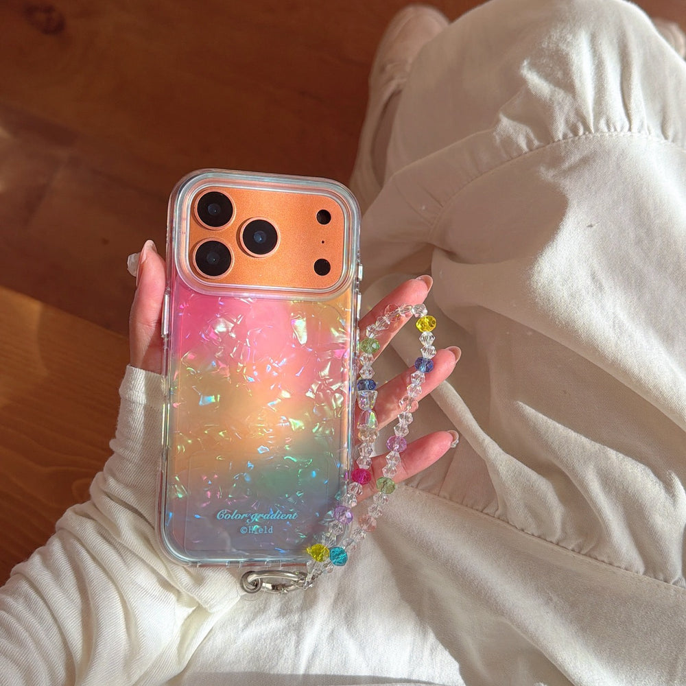 A person wearing white dress holding an orange gradient glitter iPhone case with a luxury crystal bead hand chain in soft sunlight.