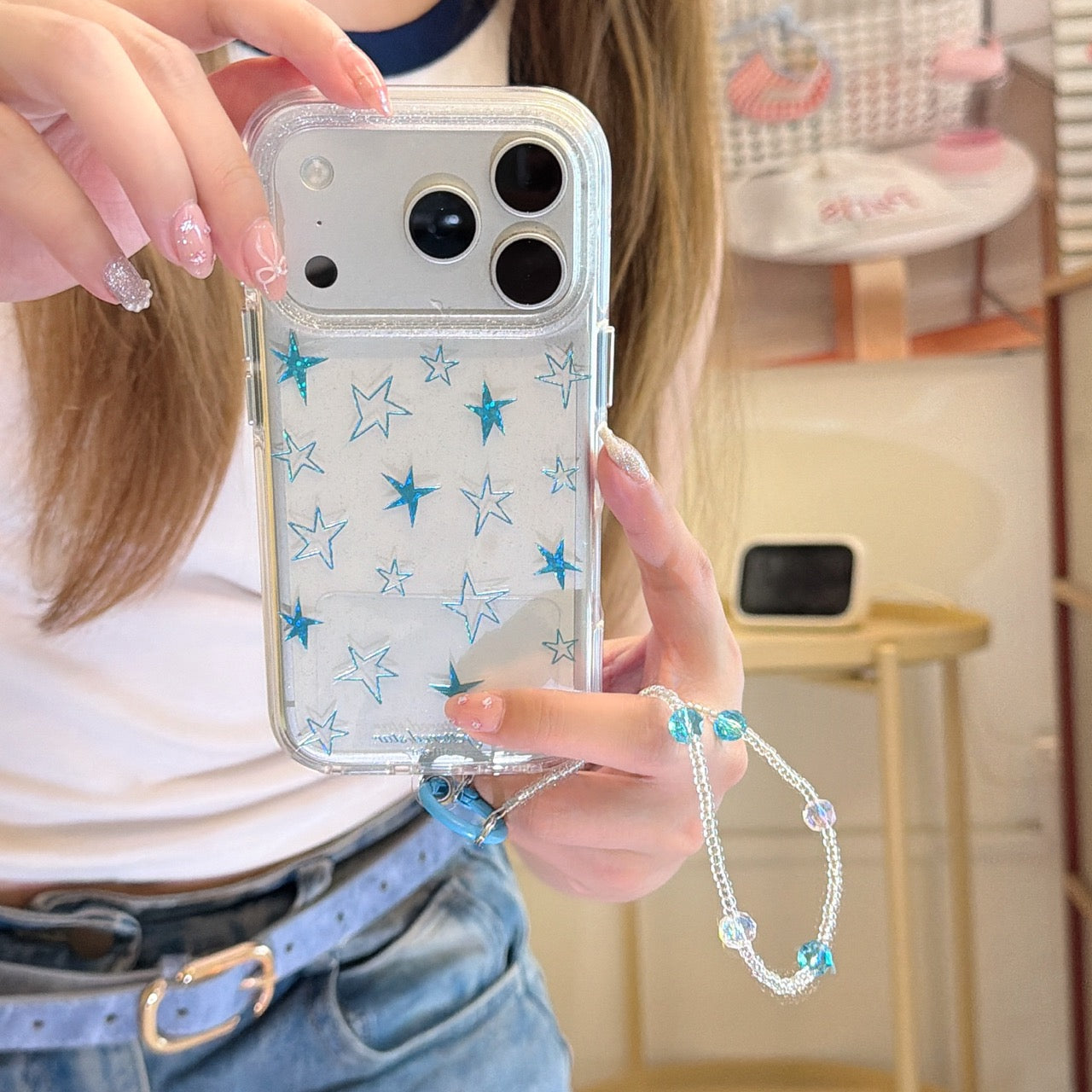 A mirror selfie showing a clear Y2K phone case with blue metallic stars and a beaded wrist strap.
