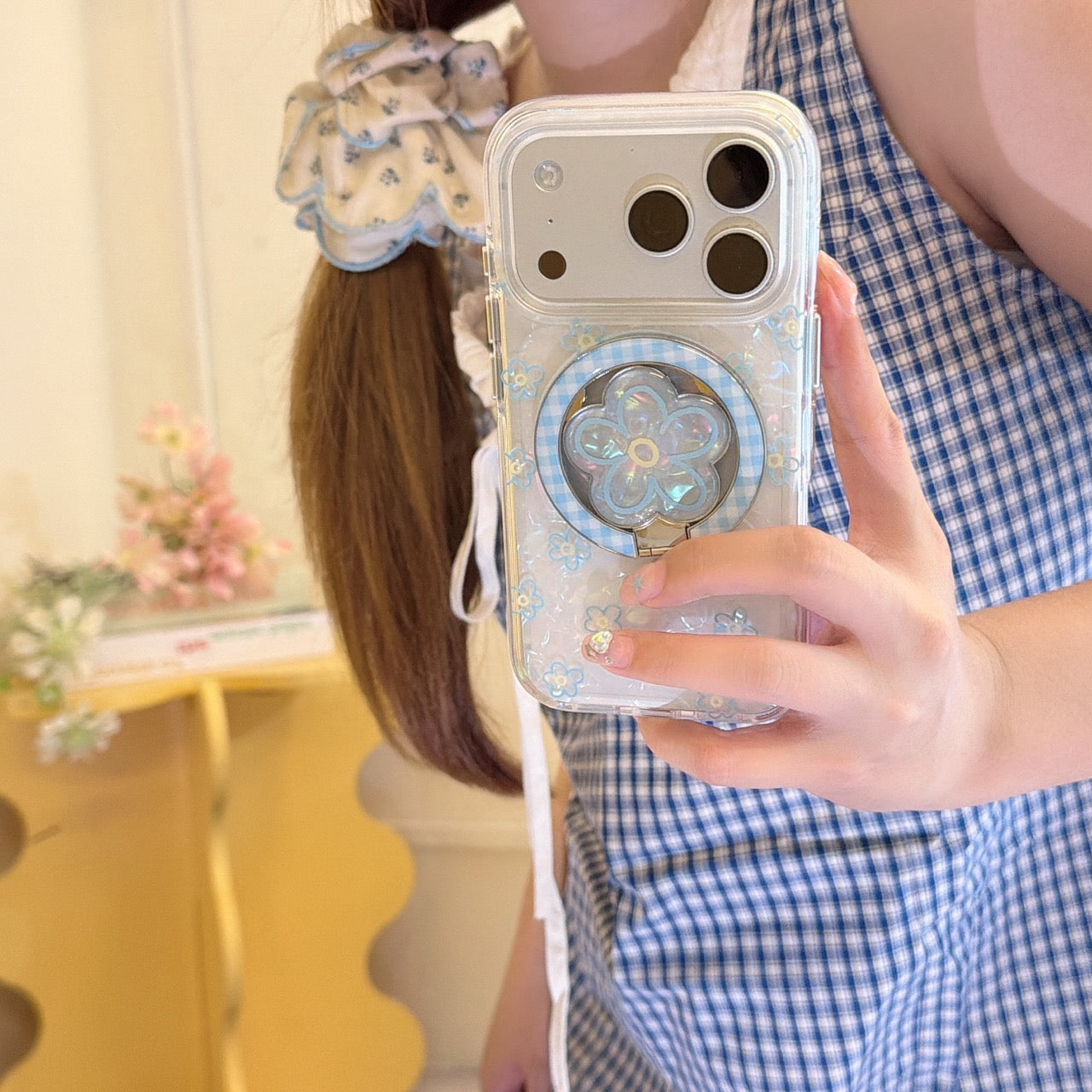 A girl in a blue gingham dress taking a mirror selfie with a pearl-texture iPhone case featuring blue flowers and a matching floral grip stand.