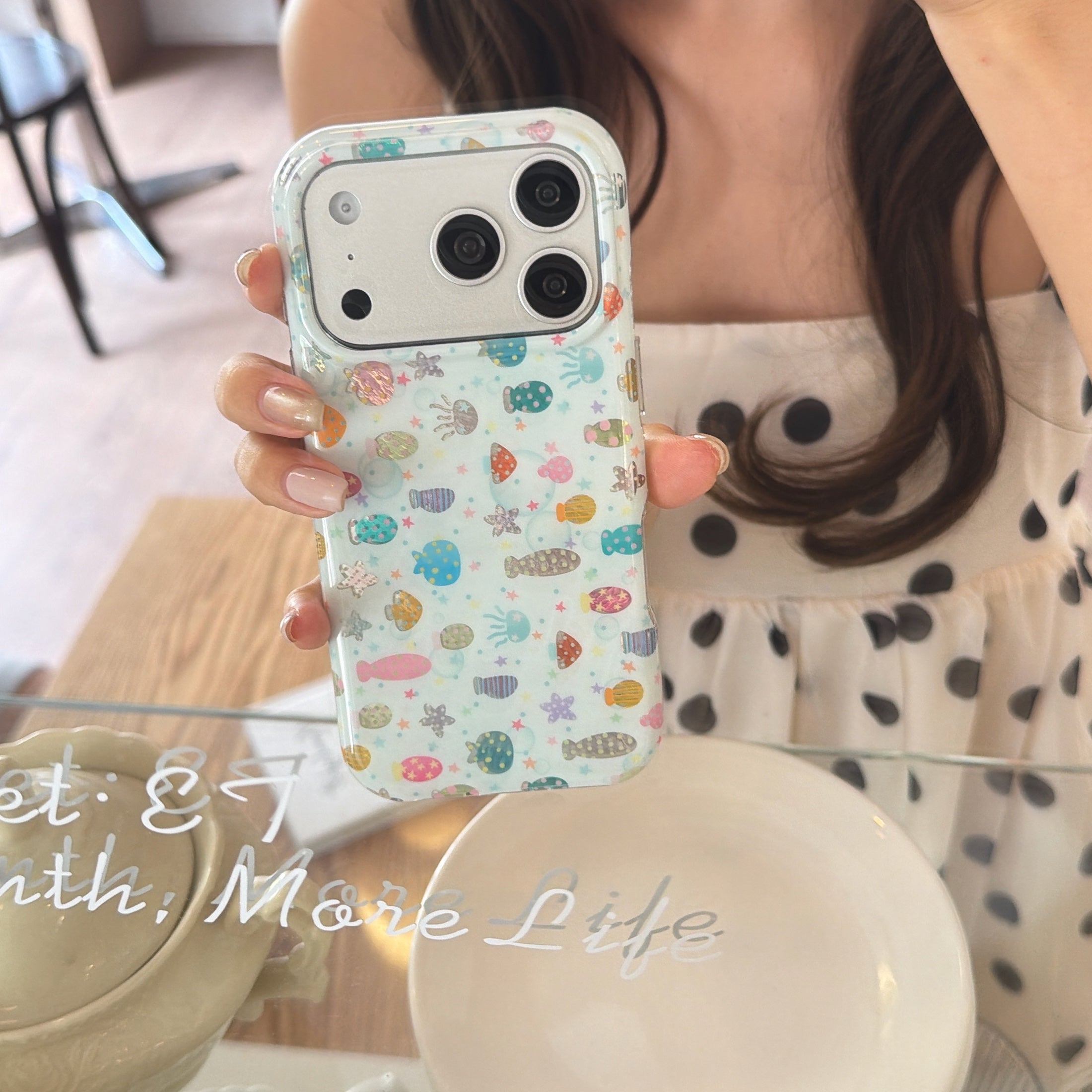 A woman holding a y2k pastel marine fish iPhone case in front of a mirrored reflection at a cafe table.