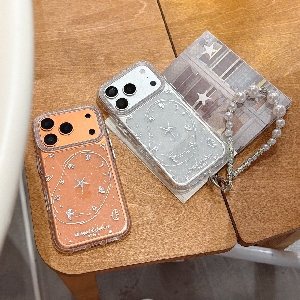 An orange and a silver glitter phone cover featuring Y2K starry sky line art and a sparkling pearl chain bracelet placed on a wooden stool.