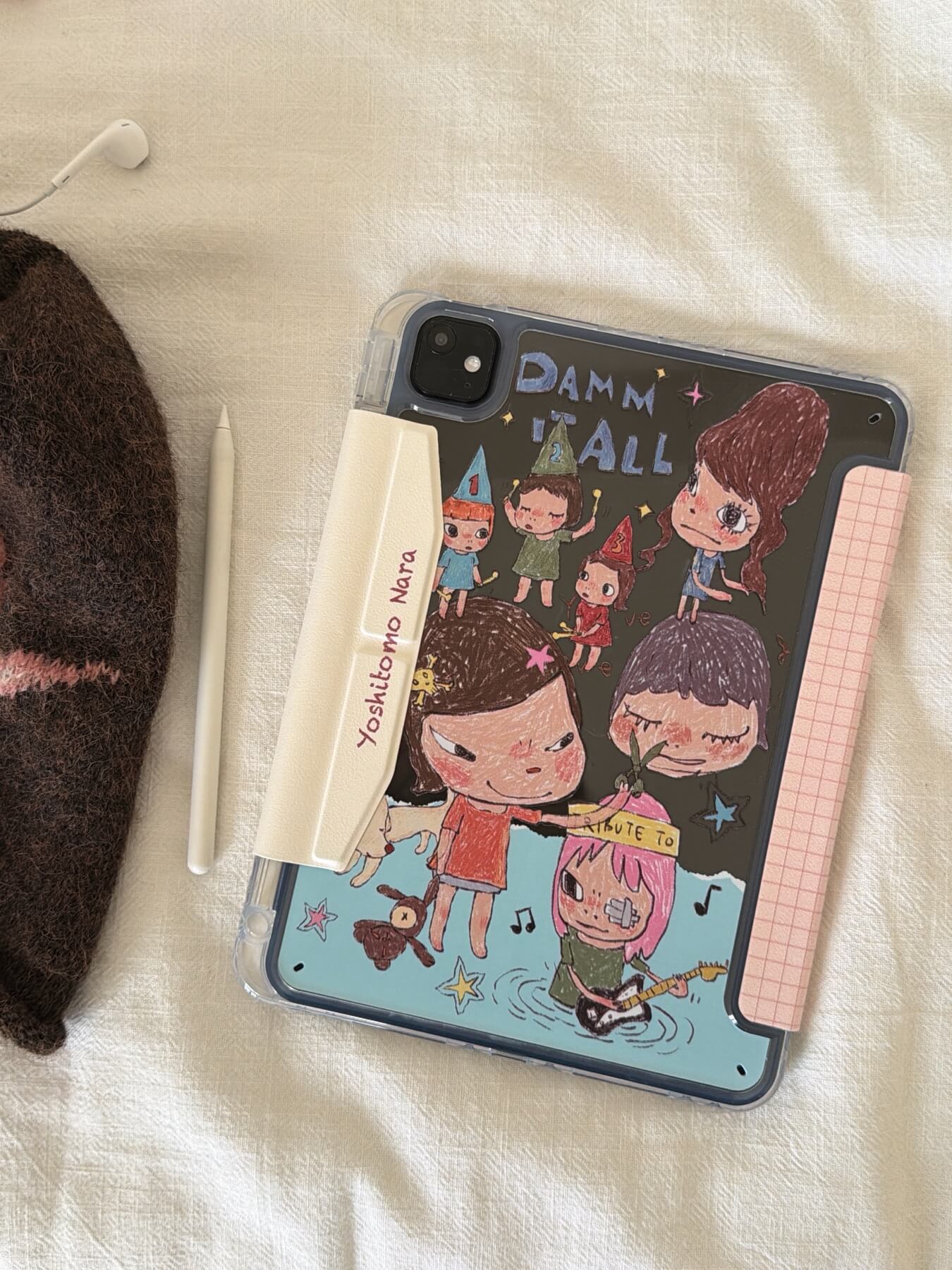 Flat lay view of a Yoshitomo Nara iPad case clear back shell with black cartoon print, an Apple Pencil, and a brown plush star beanie on a white fabric surface.