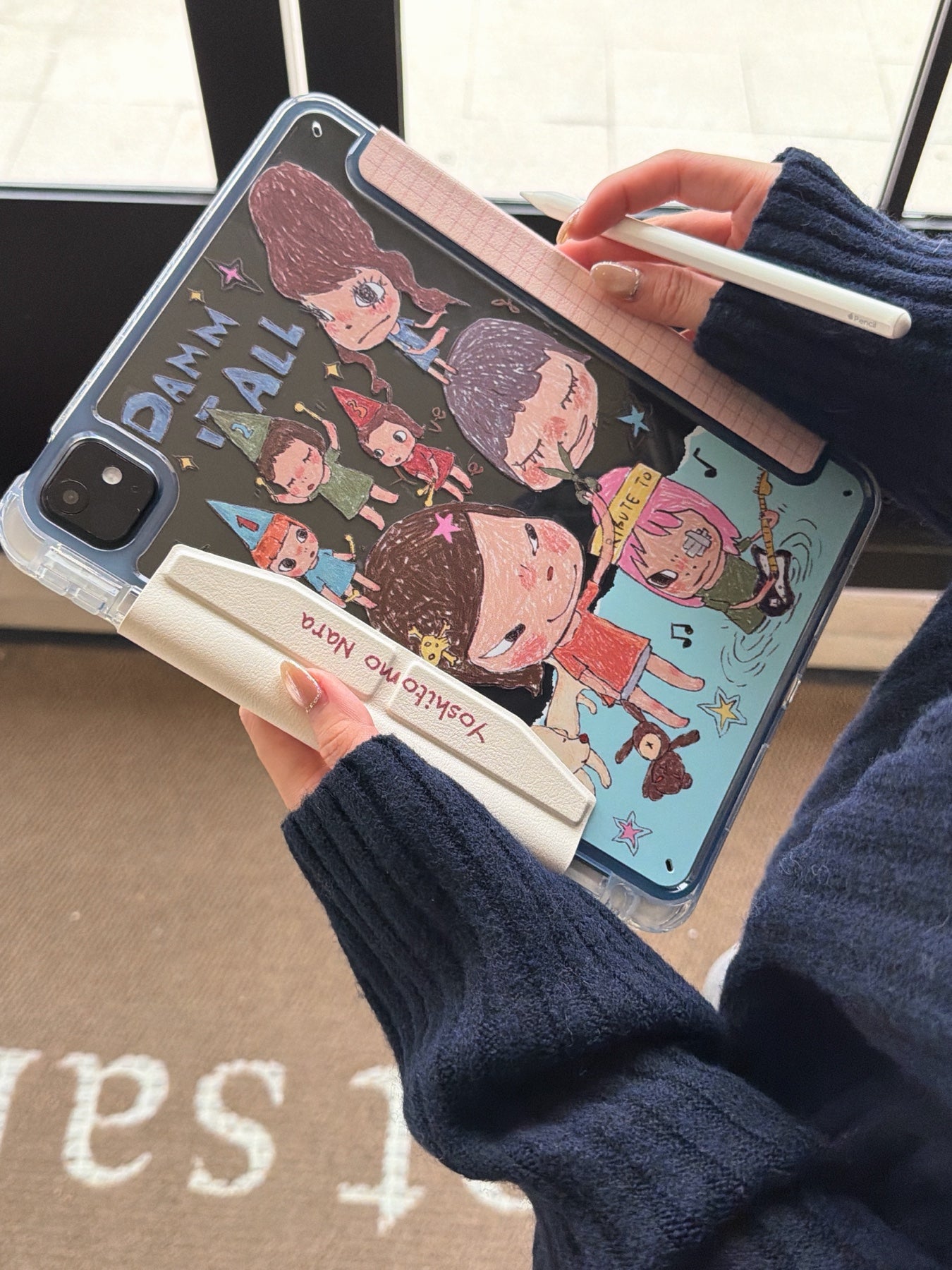 Back side of a Yoshitomo Nara iPad case with a clear crystal shell showing dark-themed artistic cartoon girls and a white protective flap closure.