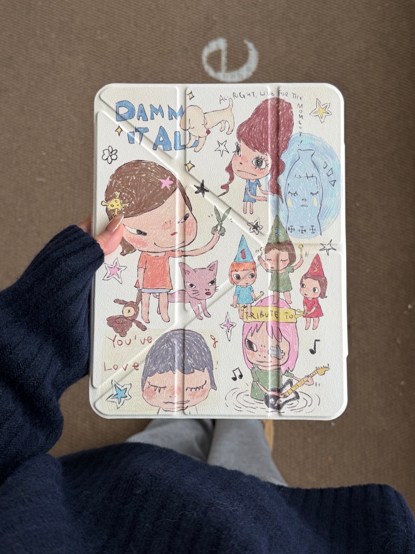 A person wearing a dark navy knit sweater holding a Yoshitomo Nara inspired iPad case vertically, displaying the cream-colored front cover with whimsical hand-drawn cartoon girls and doodles.