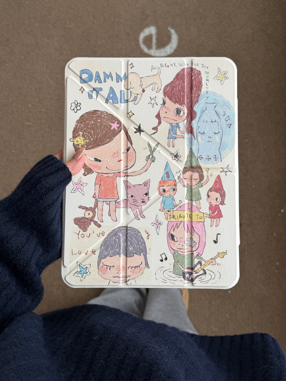 A person wearing a dark navy knit sweater holding a Yoshitomo Nara inspired iPad case vertically, displaying the cream-colored front cover with whimsical hand-drawn cartoon girls and doodles.