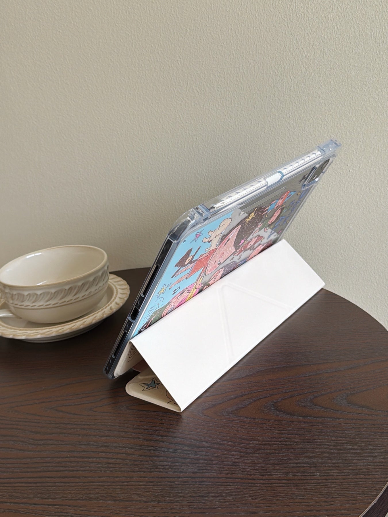 White trifold smart cover of a Yoshitomo Nara iPad case folded into a stable landscape viewing stand on a dark wooden coffee table beside a vintage white teacup.