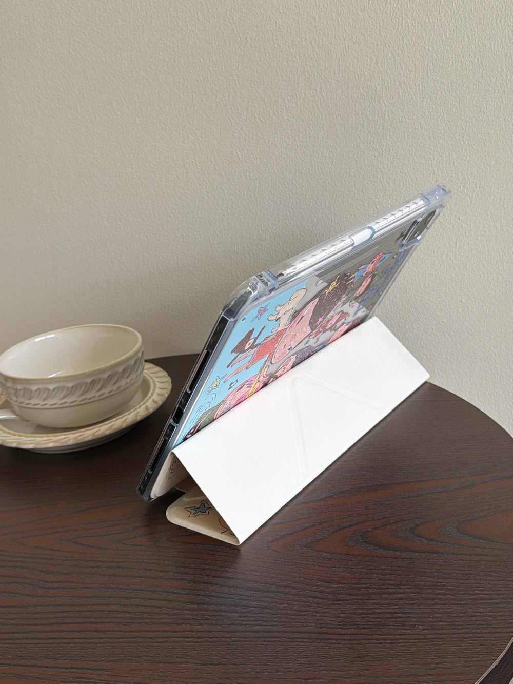 White trifold smart cover of a Yoshitomo Nara iPad case folded into a stable landscape viewing stand on a dark wooden coffee table beside a vintage white teacup.