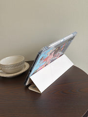White trifold smart cover of a Yoshitomo Nara iPad case folded into a stable landscape viewing stand on a dark wooden coffee table beside a vintage white teacup.