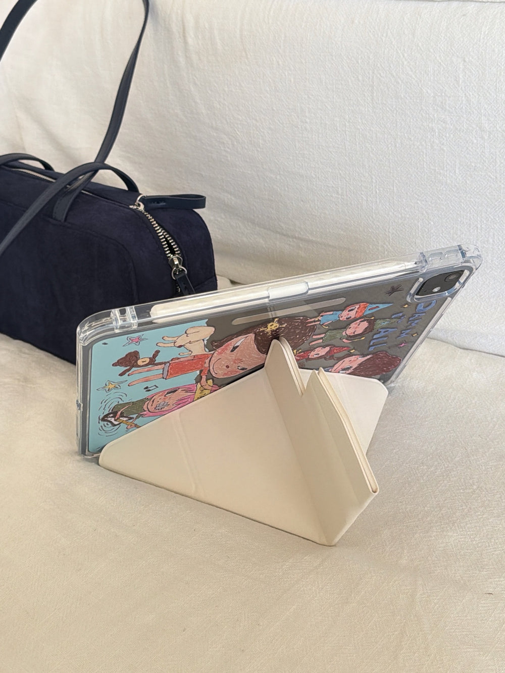 Aesthetic lifestyle shot of a cute Yoshitomo Nara iPad case propped up in an origami Y-shape stand mode on a white couch next to a dark navy blue suede handbag.