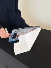 Yoshitomo Nara iPad case with clear shockproof back and white trifold smart cover standing vertically on a dark table, showing functional kickstand mode.